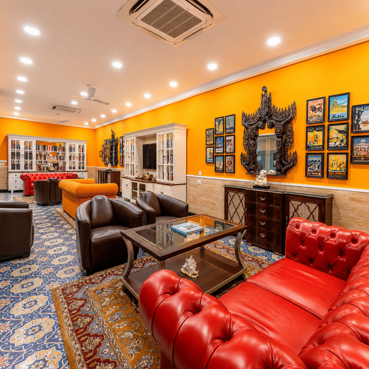 A lounge area at MAYFAIR Palm Beach Resort, Gopalpur-on-Sea. Sofas and armchairs surround a central coffee table on a patterned carpet. Framed pictures hang on the wall, and a cabinet sits in the background with lot of books in the shelves.