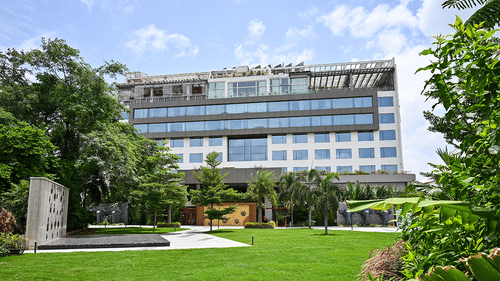 The exterior of Hotel Daspalla, a modern building surrounded by lush green lawns and trees under a blue sky.