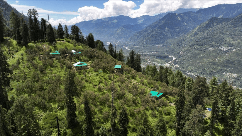 A wide-angle shot featuring several cottages scattered around on a lush green hilly area with a beautiful backdrop of the Himalayan ranges at Amara Upepo - The Sky Villa, Manali.