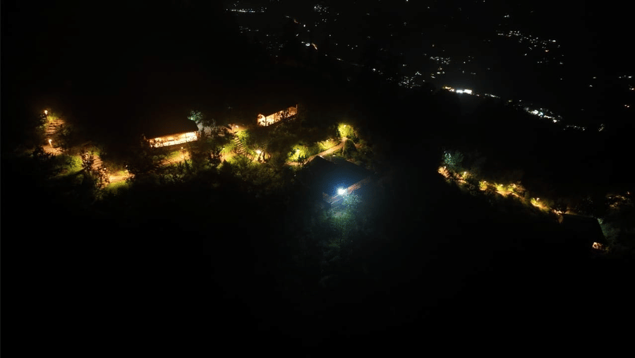 An aerial view of Amara Upepo - The Sky Village in Manali featuring various cottages being connected by a string created by the exterior pathway lights in the darkness. 