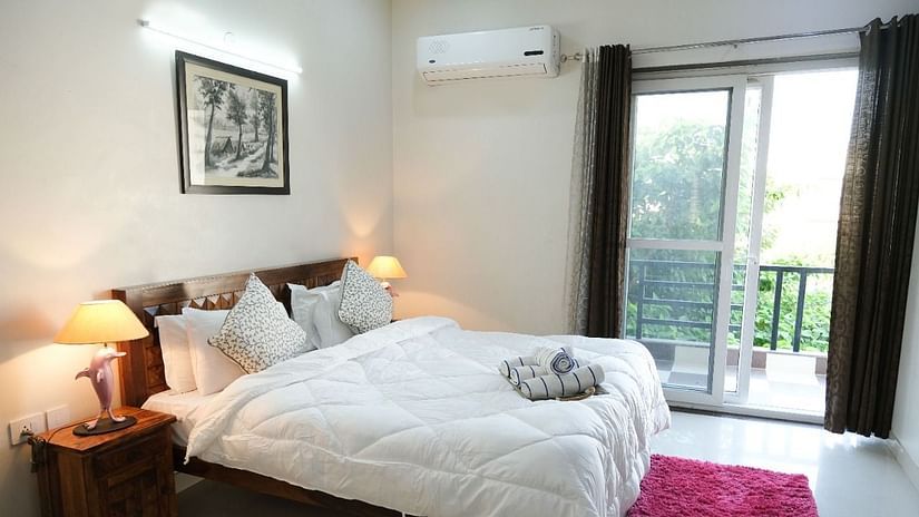 An image of a bedroom featuring a bed, a bedside table with lamp, anda a glass door leading to the balcony - LIme Tree 3BHK Serviced Apartments Near Medanta HospitalAn image of a bedroom featuring a bed, a bedside table with lamp, anda a glass door leading