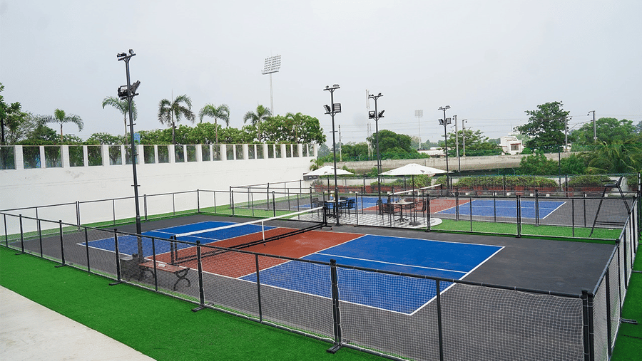 A fenced outdoor sports court with marked playing surfaces, netted boundaries, lighting poles, and surrounding greenery at Clarks Avadh, Lucknow.