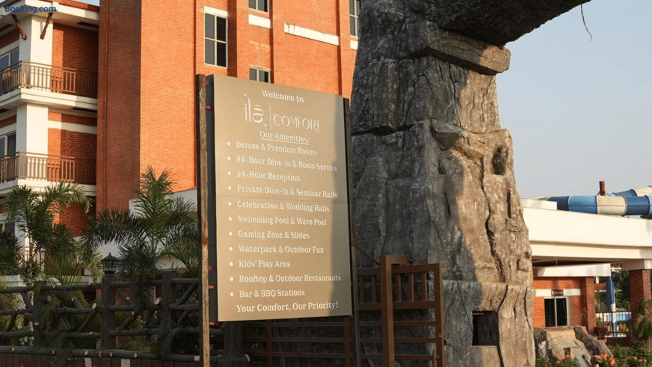 Amenities signboard by the gate listing rooms, dining, pools and play areas, with the red-brick building and water slide behind, at Ila Comfort, Harkapur.