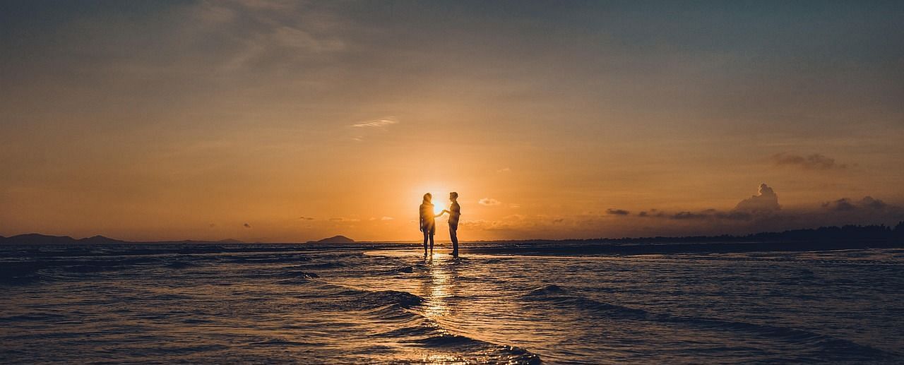 A couple standing on the shoreline during sunset and their reflection visible in the ocean
