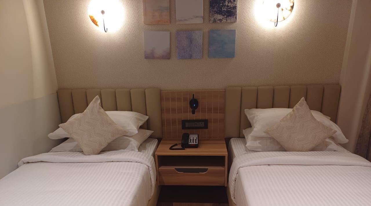 Premium Room at Hotel Paai, Hosur featuring twin beds placed at two corners of the room. Above each bed there are lights on the wall. Between the beds there is a bedside table with a telephone and socket. 