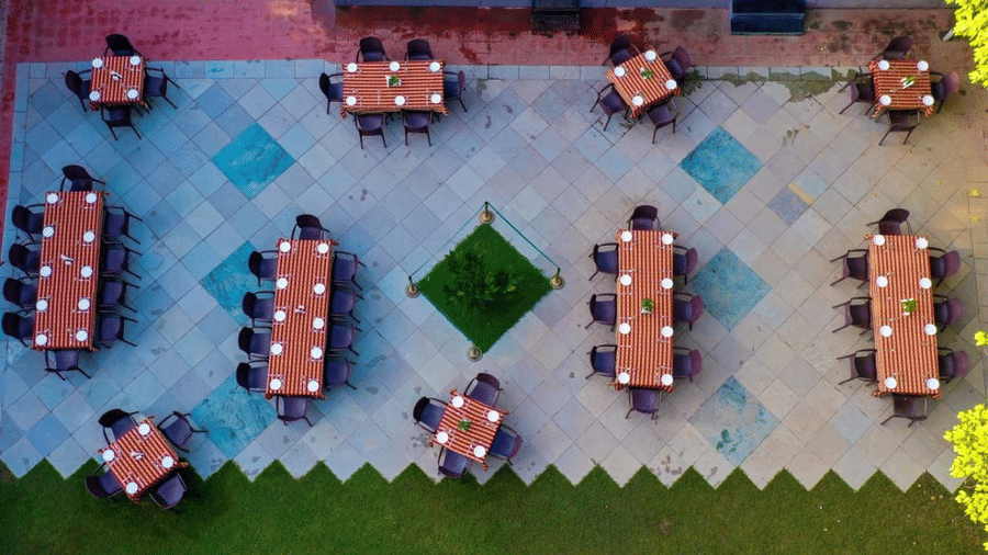 A top-down view shows multiple long dining tables arranged on a patterned outdoor patio next to a green lawn at Amantra Shilpi Resort, Udaipur.