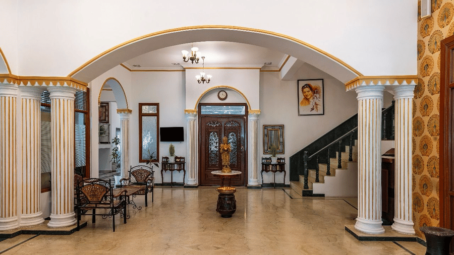 A spacious tiled lobby area with white archways, pillars, seating, and a staircase, at Amantra Shilpi Resort, Udaipur.