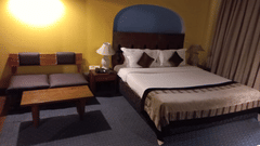 Guest room at The Bristol Hotel, Gurgaon with a king-size bed, an arched headboard, striped bedspread, brown cushions, a bedside lamp, and a cushioned bench.