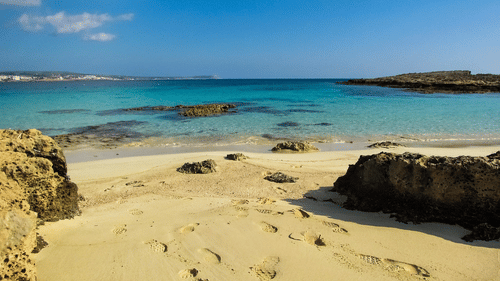 A sandy beach with shallow turquoise water, rocky formations along the shoreline, and a clear sky, showing a quiet coastal landscape by the sea.