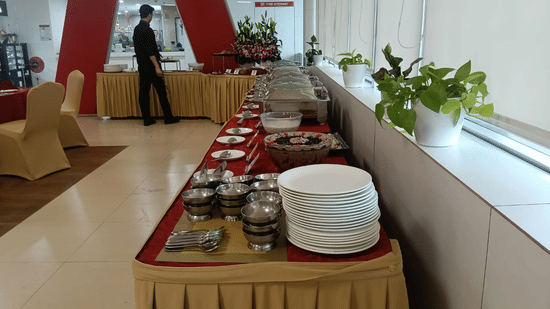 A catering setup inside a room. Tables are arranged in a row with tablecloths and skirting. Plates, bowls, and serving dishes are positioned on top of the tables. A person stands in the background near a window, and plants sit on the window sill.