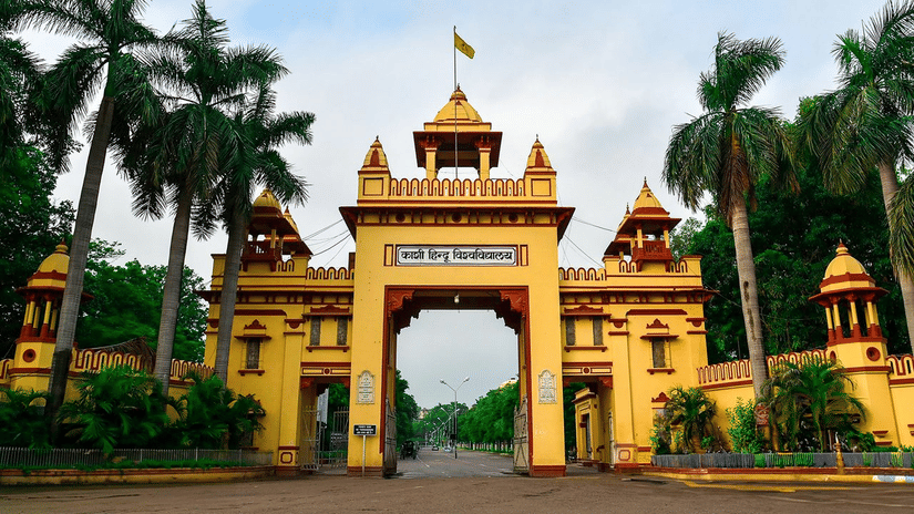 The grand entrance gate of Banaras Hindu University features traditional arched doorways, decorative domes, and tall surrounding palm trees.