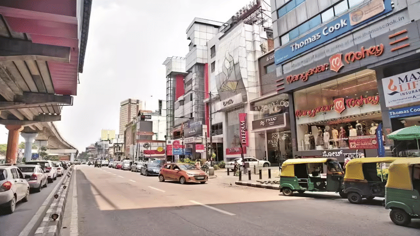 Vehicles and auto-rickshaws drive past bustling commercial buildings and retail storefronts along the wide street of MG Road during daytime.