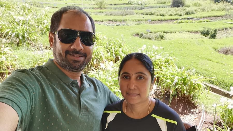 Man and woman posing for a selfie with lush green fields and garden plants in the background.