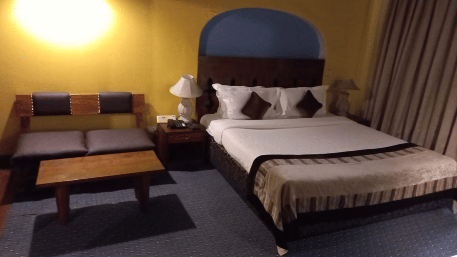 Guest room at The Bristol Hotel, Gurgaon with a king-size bed, an arched headboard, striped bedspread, brown cushions, a bedside lamp, and a cushioned bench.