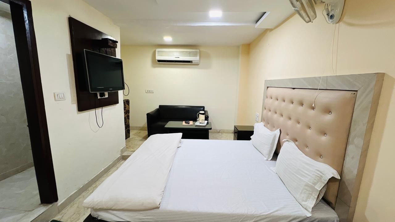 A clean and simple deluxe room with a comfortable bed, a small sofa, and a wall-mounted TV, offering basic comforts. | Deluxe Room | Ajay Guest House
