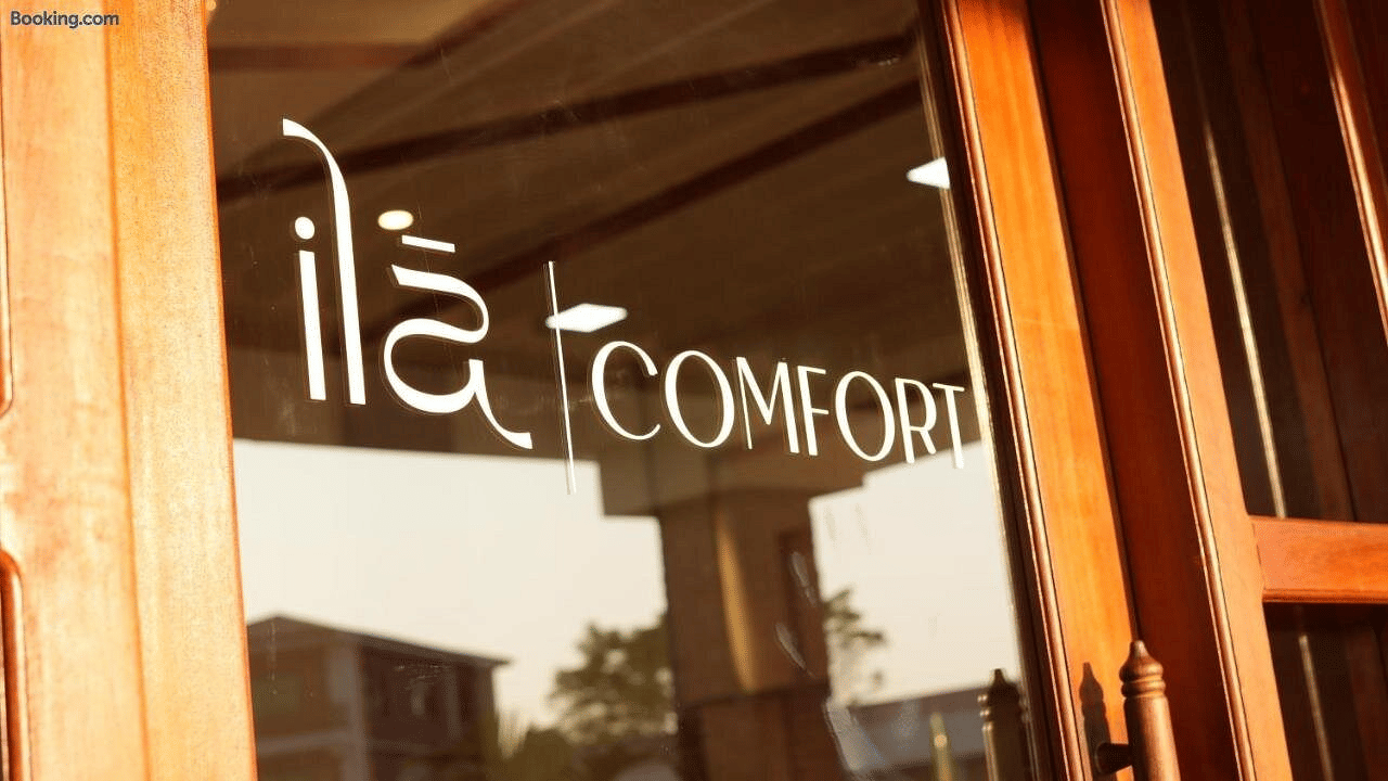 Brand signage etched on the wood-framed glass entrance door, with the porch and greenery reflected in the glass, at Ila Comfort, Harkapur.