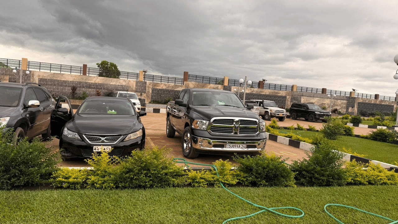 A wide shot of a modern building's exterior with two parked black SUVs, a lawn area, and a cloudy sky at Luffu Club.