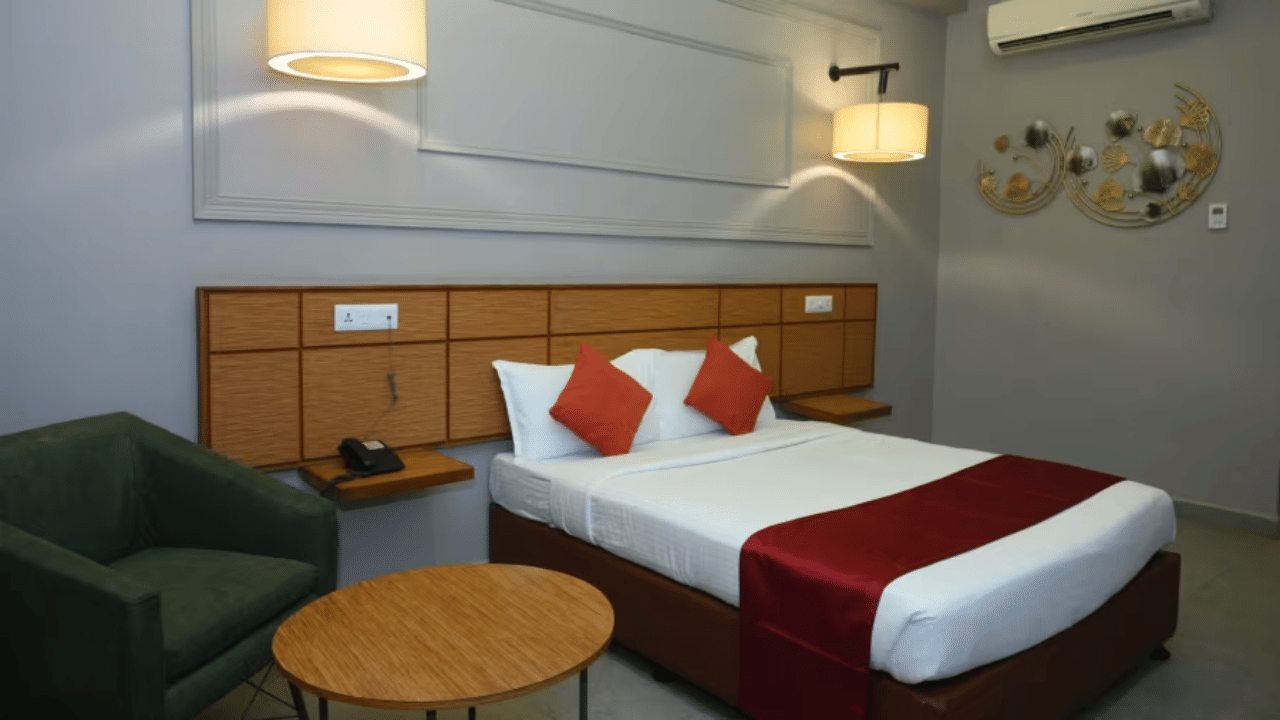 Bedroom with double bed, red runner, pillows, green chair, table and wall lamps at Max Hotel Prayagraj.