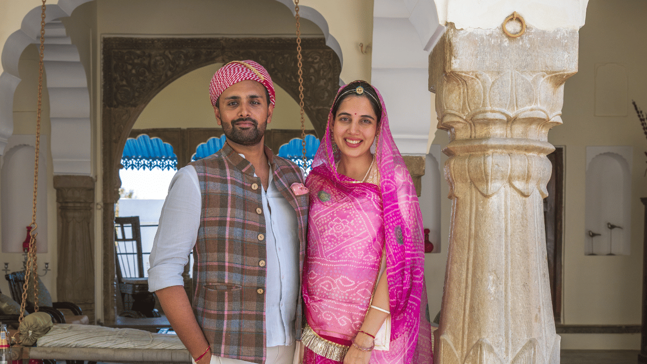 Divyaraj and Nityasri in traditional Indian wedding attire standing inside a heritage palace, with carved arches and pillars in the background.