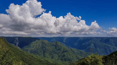 A beautiful landscape of Shillong featuring lush green mountains with a bright blue sky and distinctive clouds floating over it.
