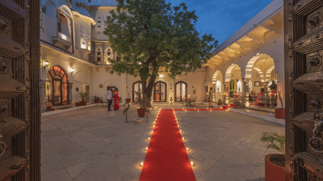 Evening view of a heritage palace courtyard with a red carpet lined with oil lamps leading towards a large tree, surrounded by warmly lit arched facades and guests mingling under the twilight sky.