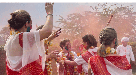Group of women in white and red sarees joyfully celebrating Holi, throwing colourful powder in the air during a vibrant outdoor festival scene.