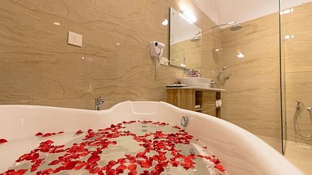Bathtub filled with water and red rose petals in a modern bathroom featuring beige tiled walls, a vanity mirror, and a walk-in shower.