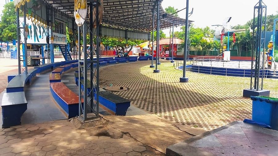 An amusement park stage with a blue walkway, surrounded by palm trees and fairground booths under a partial roof.