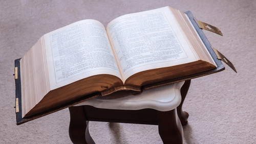 An open book placed on a wooden stand with a pen resting on the pages on a table.
