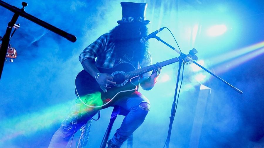 Slash, silhouetted in a top hat and sunglasses, energetically plays guitar on a smoky, brightly lit stage.