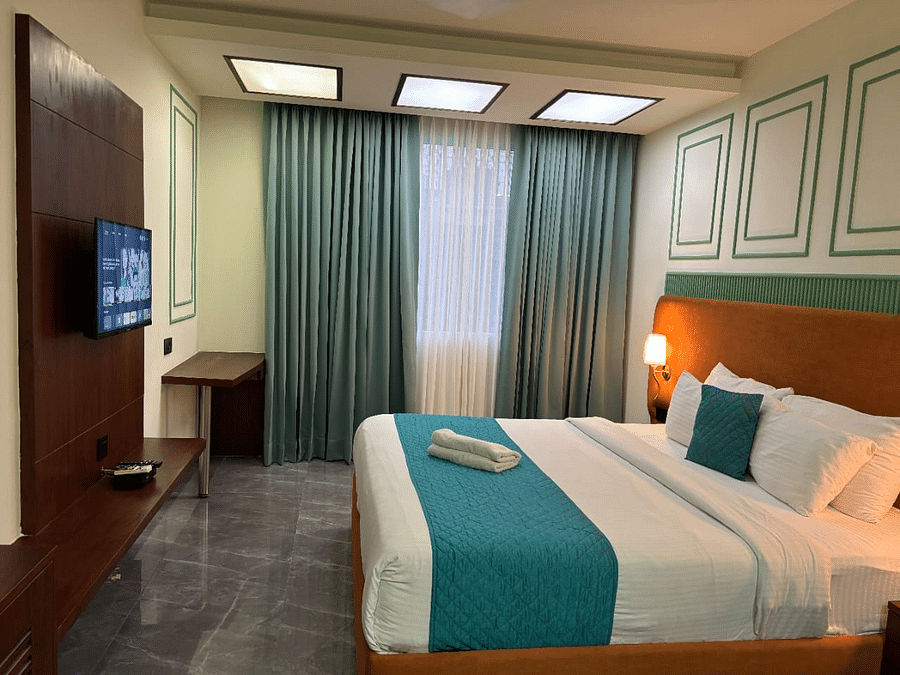An interior view of a hotel room featuring a neatly made bed with pillows , a television and a window with curtains -  Asian Suites IFFCO Chowk Metro Station, Gurgaon.