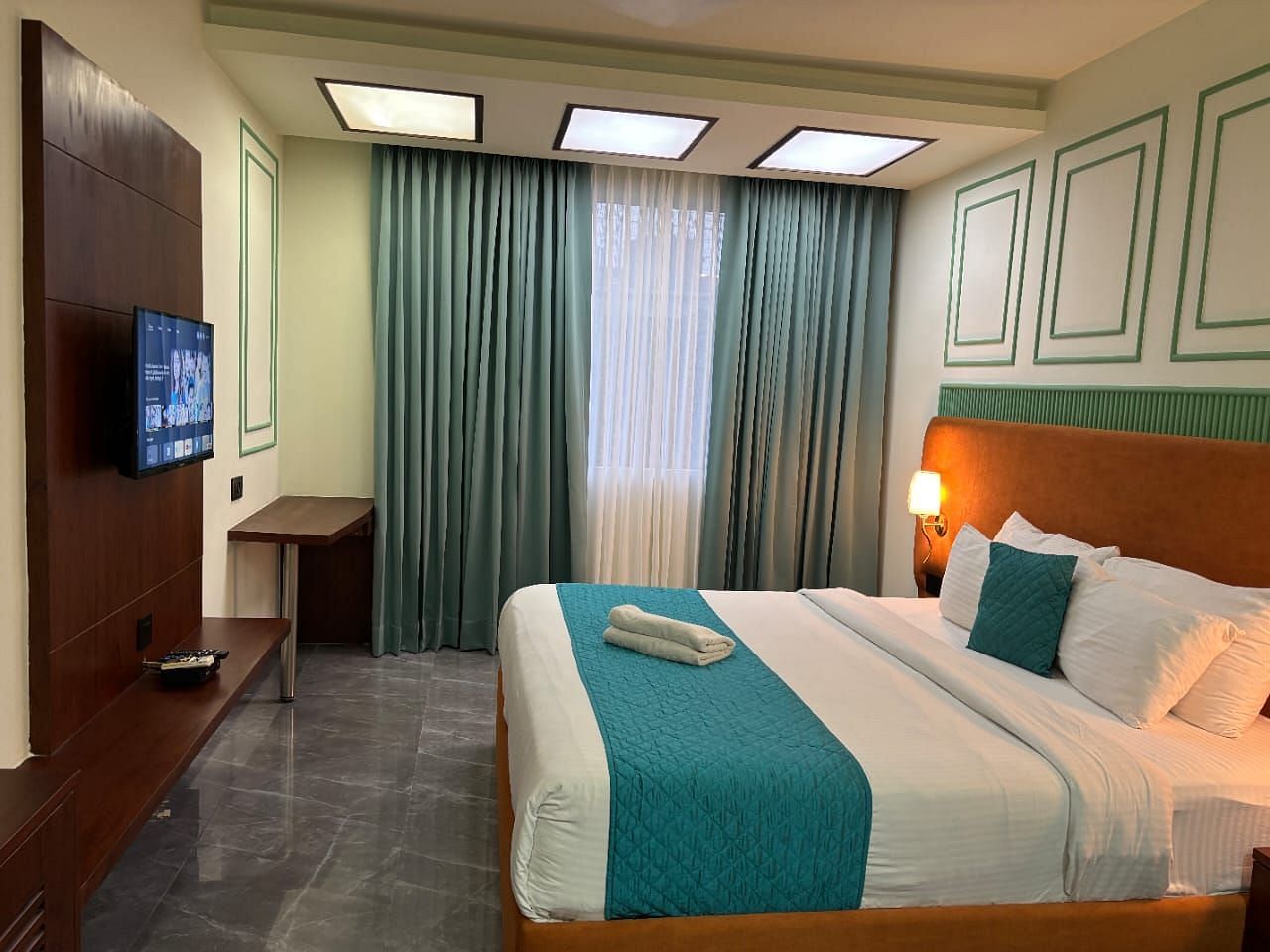 A comfortable room showing a double bed with blue accents, a television, and curtains - Asian Suites IFFCO Metro, Gurgaon