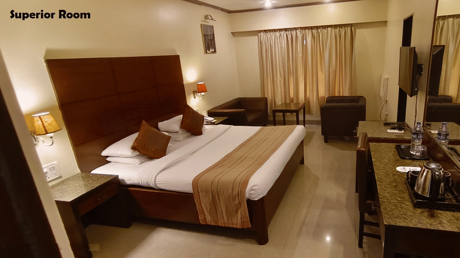 A Superior Room featuring a bed with a runner, a wood panelled headboard, 2 sofas facing a coffee table, and a big mirror on the wall | Kings International Juhu