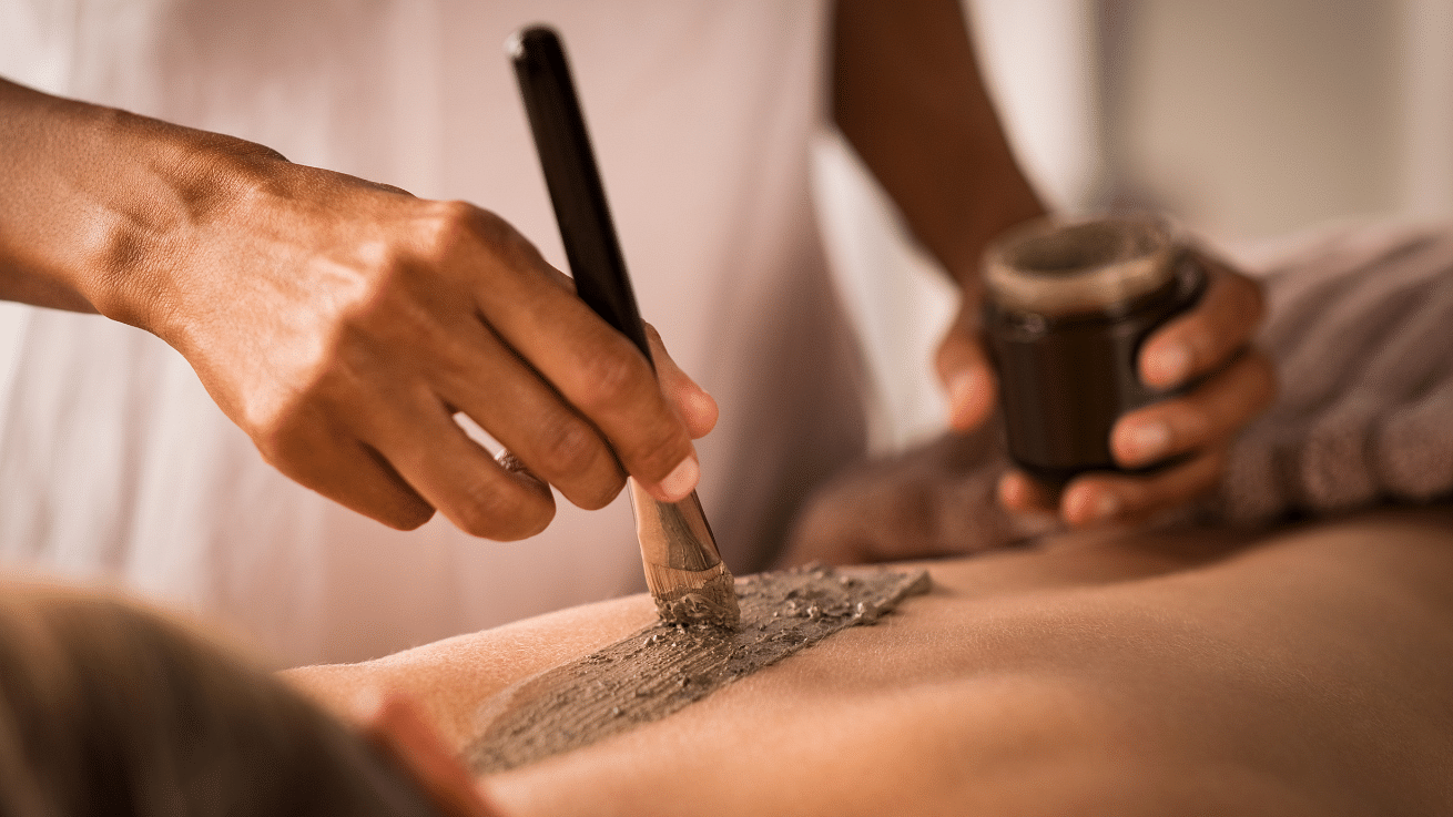 A person receiving a body scrub treatment, with a close-up of hands applying the scrub on their back using a brush.