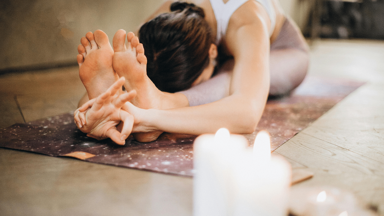 A person performing a seated forward bend yoga pose on a mat, with their head touching their knees and hands reaching for their feet, surrounded by lit candles.
