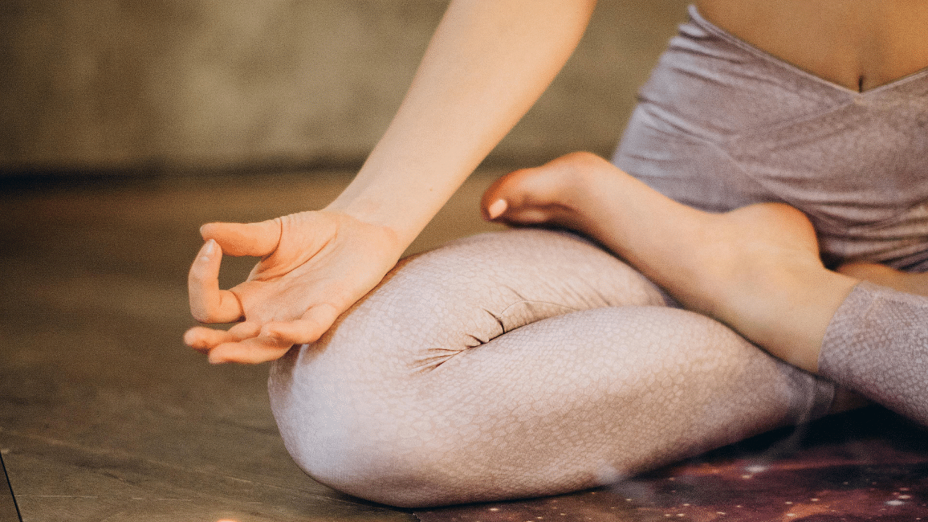 Close-up of a person in a seated lotus yoga pose, wearing light-colored leggings, with one hand resting on the knee in a mudra position.