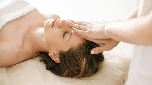 Gentle hand touching a woman's forehead as she relaxes on a white surface.