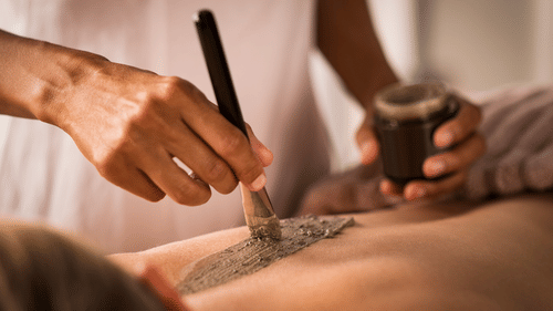 Hands applying a gray clay mask with a brush on someone's back in a spa treatment.