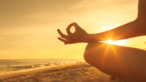 A hand in a yoga mudra with the sun setting through the fingers on a beach.