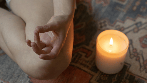 A close-up of a meditative hand gesture next to a lit candle on a patterned rug.