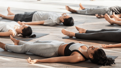 People in a yoga class lying down in relaxation pose, in a serene, bright room.