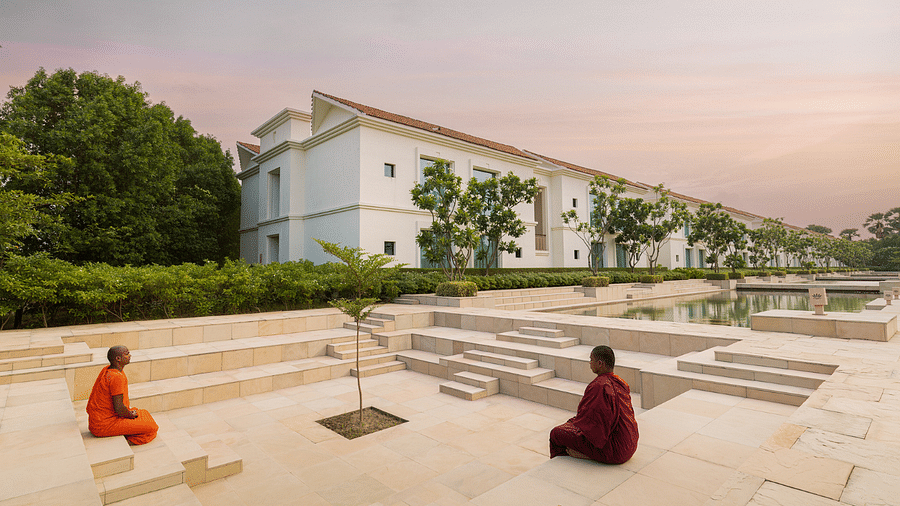 A serene scene with two monks seated in meditation, facing a large reflective pool with a modern building in the background, framed by trees and a pinkish sky.