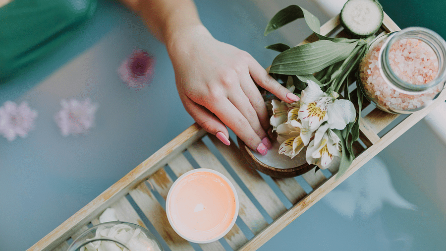 A person reaching for a flower on a wooden bath tray with a lit candle, salt jar, and cucumbers.