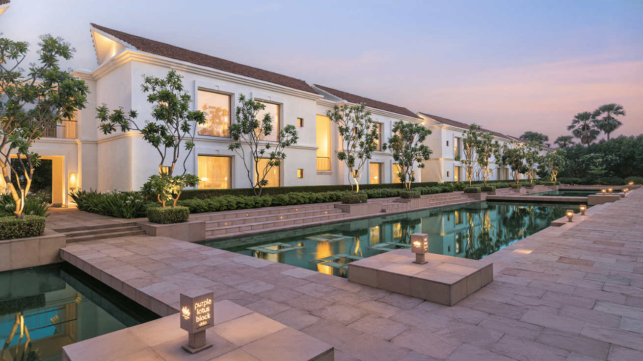 The exterior of a modern, luxurious building at dusk, with lighting illuminating the structure and a calm reflective pool in the foreground.