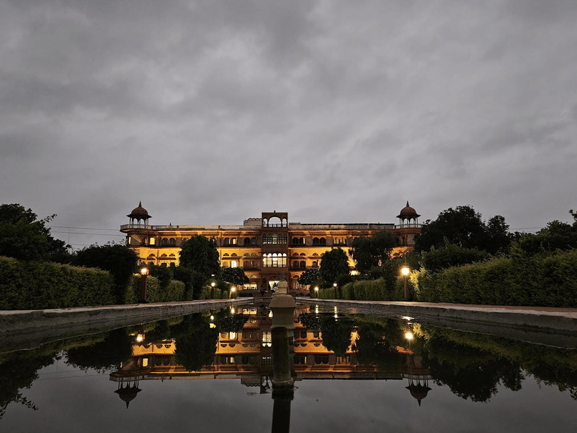 A well lit facade of Umaid Palace, a premiere resort near Bhangarh Fort, featuring stretched and multi storey building with lights on. The property is covered by trees and there is also a huge fountain in the front yard.