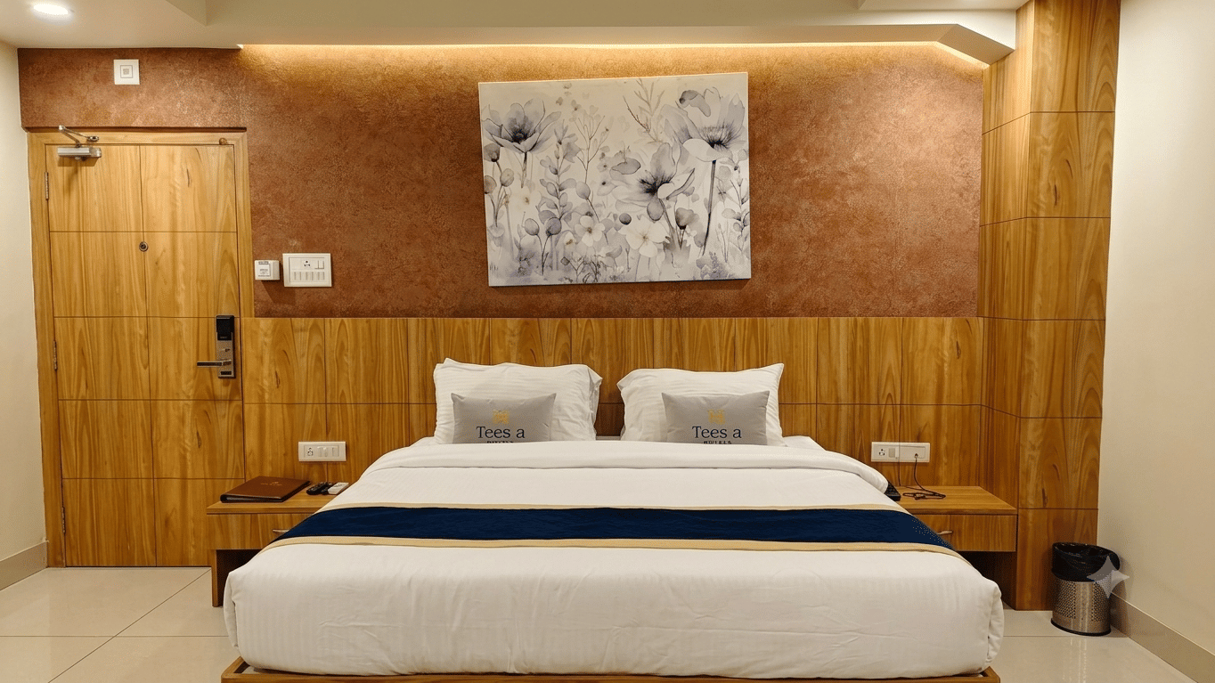 Deluxe Room at Teesta Stays, Rampurhat showing a double bed positioned in front of a wooden headboard, wall painting, room entrance door, twin side tables, and a bin placed on the floor tile.