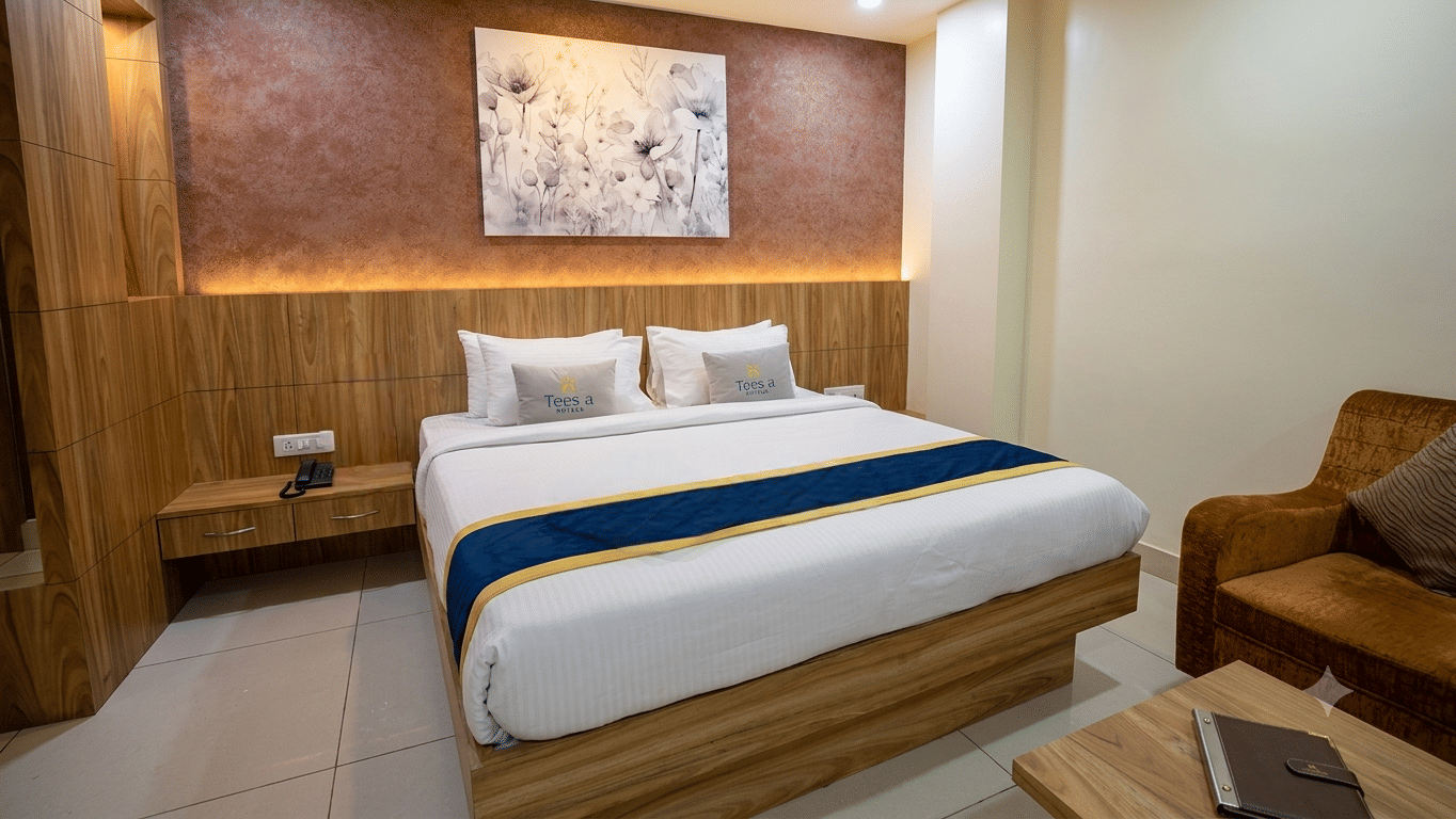 Superior Room featuring a double bed with pillows and runner at Teesta Stays, Rampurhat. It is also showing a wooden headboard, side table, artwork mounted on the wall panel, a wooden sofa chair, and a table with an open notebook.