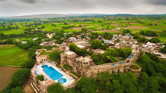 drone shot of Hill Fort Kesroli 14th Century Alwar where swimming pool and multiple structures are surrounded by lush green landscape and cloudy blue sky in the background.