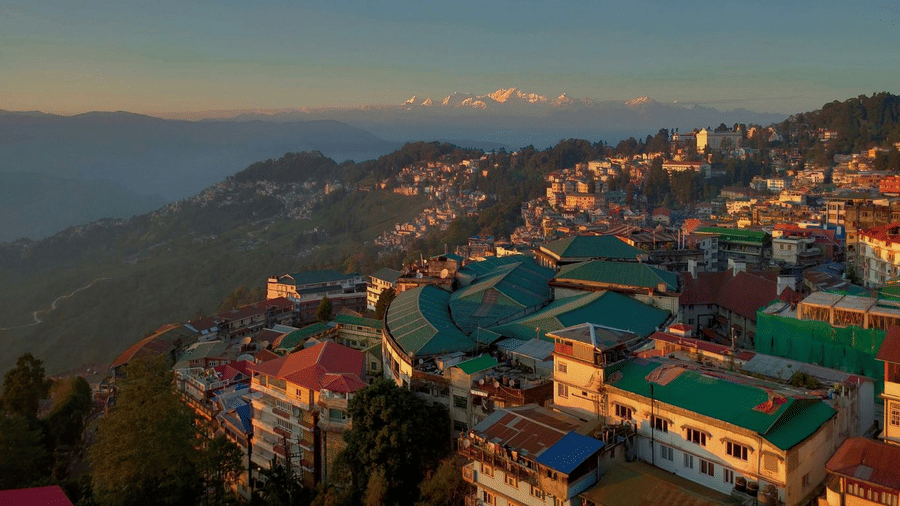 Aerial view of Darjeeling town at sunrise with colorful hillside buildings and snow-capped Kanchenjunga peaks in the distance.