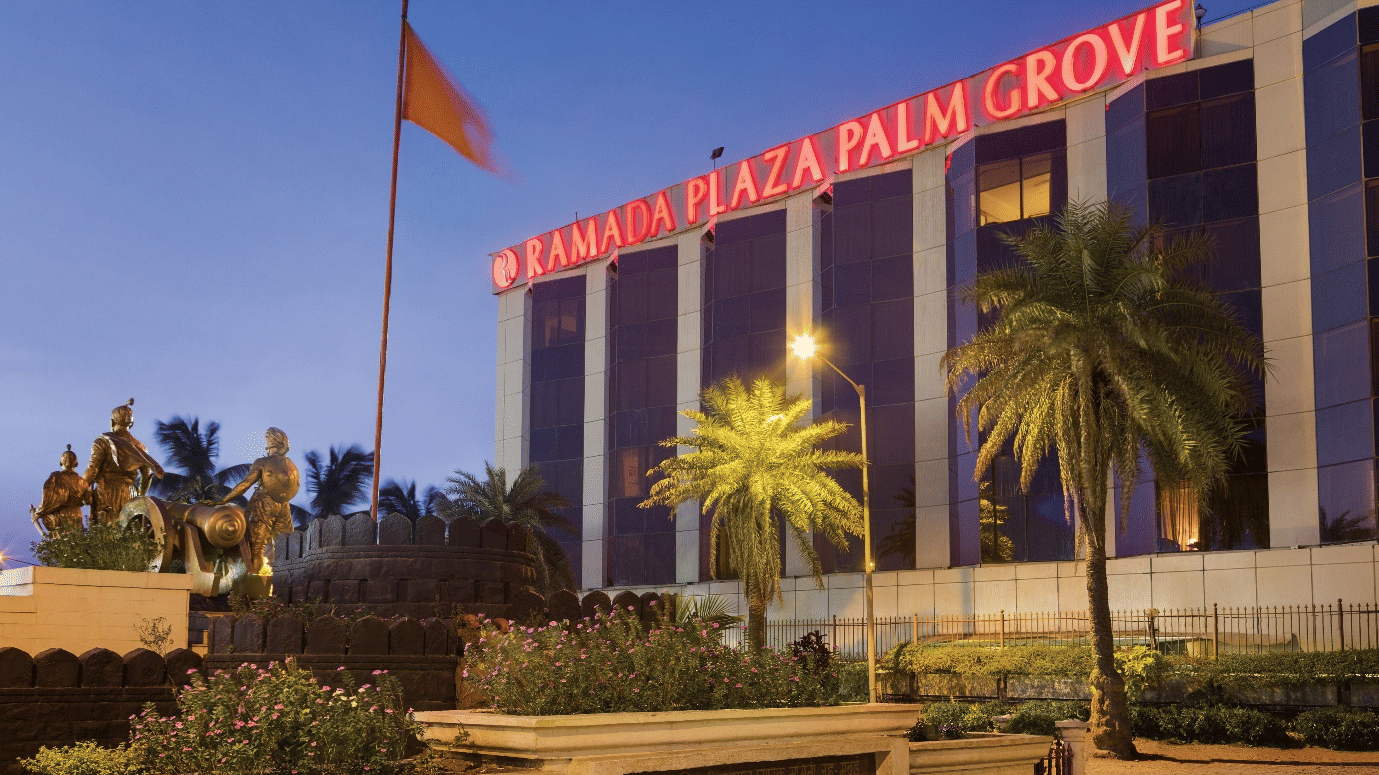 Exterior view of Ramada Plaza by Wyndham Palm Grove, showcasing its illuminated facade, palm-lined surroundings, and prominent city landmark setting.